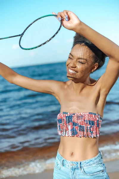 Portrait of smiling african american woman with badminton racquet on beach — Stock Photo