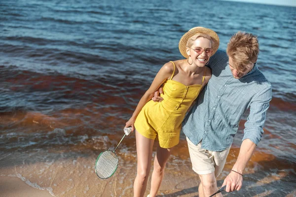 Porträt eines glücklichen Paares mit Badminton-Ausrüstung, das sich am Sandstrand mit Meer im Hintergrund umarmt — Stockfoto