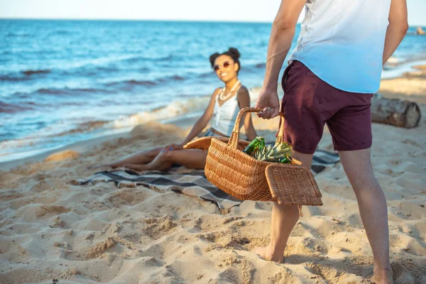 Vista parcial de pareja interracial teniendo picnic en la playa de arena - foto de stock