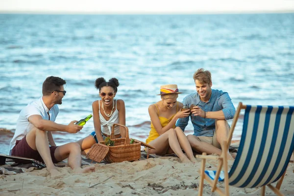 Interracial sonriendo jóvenes amigos con bebidas descansando en la playa de arena juntos en el día de verano - foto de stock