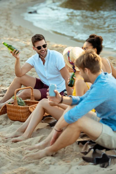 Interracial sonriendo jóvenes amigos con cerveza descansando en la playa de arena juntos en el día de verano — Stock Photo