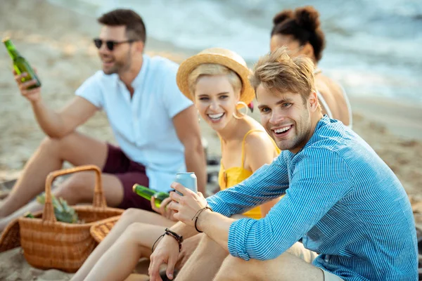 Interracial sonriendo jóvenes amigos con cerveza descansando en la playa de arena juntos en el día de verano - foto de stock