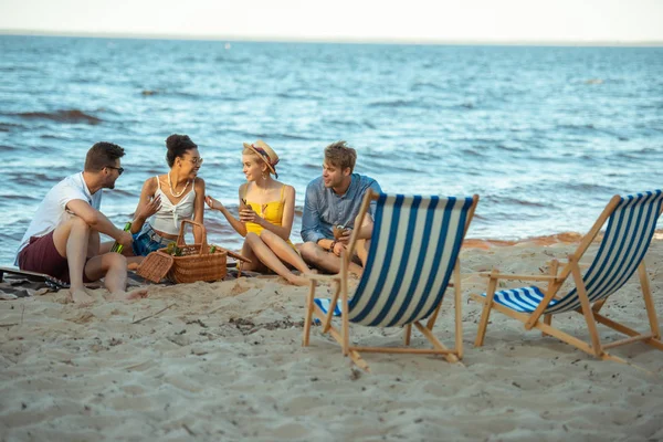 Interracial sonriendo jóvenes amigos con cerveza descansando en la playa de arena juntos en el día de verano - foto de stock