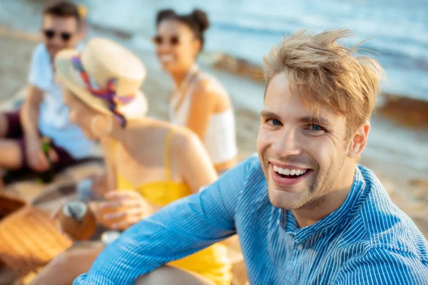 Selektiver Fokus eines fröhlichen jungen Mannes, der am Strand in die Kamera schaut, und seiner multirassischen Freunde in der Nähe — Stockfoto