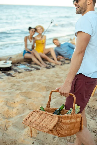 Vue partielle de l'homme avec panier de pique-nique et amis multiethniques sur la plage de sable — Photo de stock