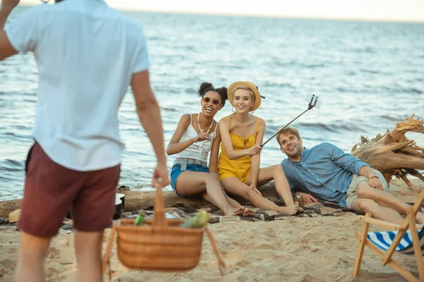 Partial view of man with picnic basket and multiethnic friends on sandy beach — Stock Photo