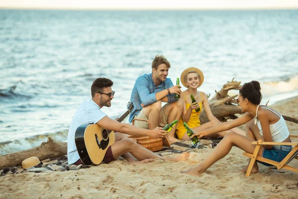 Amigos sonrientes multiétnicos con bebidas y guitarra acústica descansando en la playa juntos - foto de stock