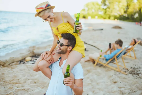 Вибірковий фокус щасливі жінка, сидячи на плечах boyfriends і друзями відпочивав позаду на пляжі — стокове фото