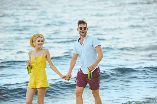 Cheerful couple with beer in glass bottles walking on beach — Stock Photo