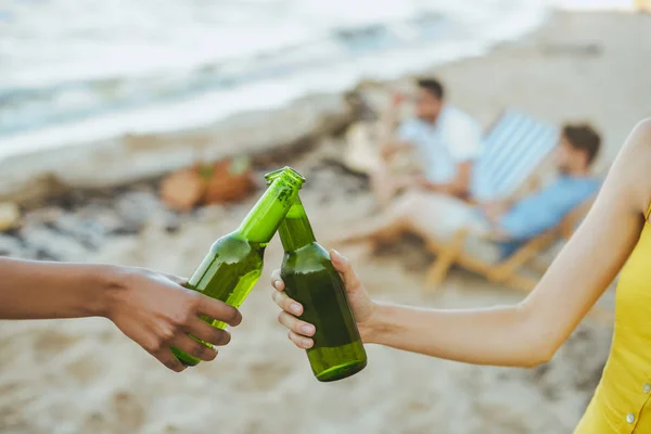 Messa a fuoco selettiva di amici multirazziali clinking bottiglie di vetro di birra mentre riposano sulla spiaggia di sabbia insieme — Foto stock