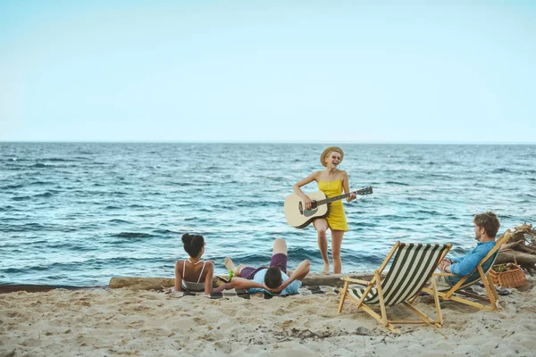Молода жінка грає на акустичній гітарі для багатоетнічних друзів на піщаному пляжі — стокове фото