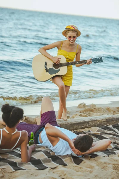 Focalizzazione selettiva della giovane donna che suona la chitarra acustica per amici multietnici sulla spiaggia sabbiosa insieme — Foto stock