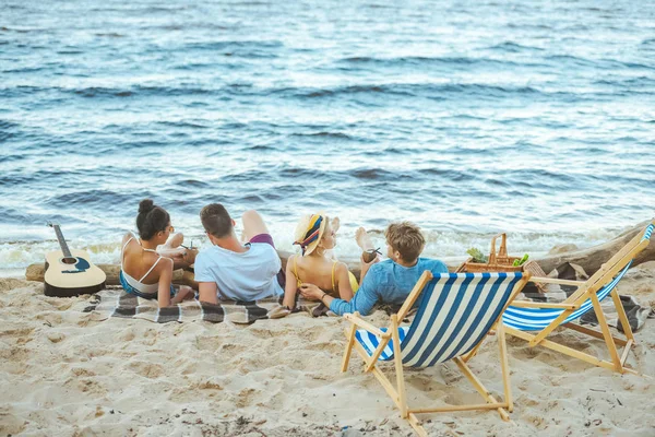 Vista trasera de amigos multiétnicos descansando sobre una manta en la playa de arena - foto de stock