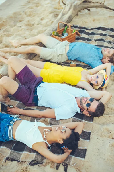 Вид сбоку на молодых людей разных рас, лежащих на одеяле, проводя время на песчаном пляже — стоковое фото