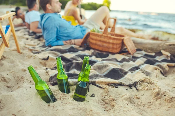 Foco seletivo de garrafas de vidro de cerveja na areia e jovens inter-raciais descansando perto de — Fotografia de Stock