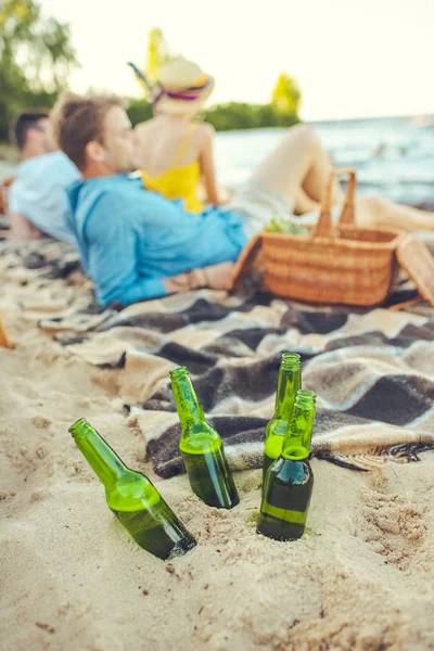 Selective focus of glass bottles of beer in sand and interracial young people resting near by — Stock Photo