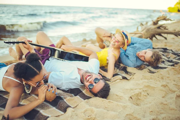 Amigos multiculturales con cocteles de coco y guitarra acústica descansando en la playa de arena - foto de stock