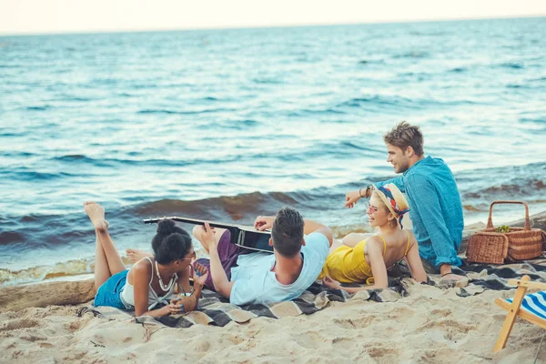Мультикультурные друзья с кокосовыми коктейлями и акустической гитарой отдыхающие на песчаном пляже — стоковое фото