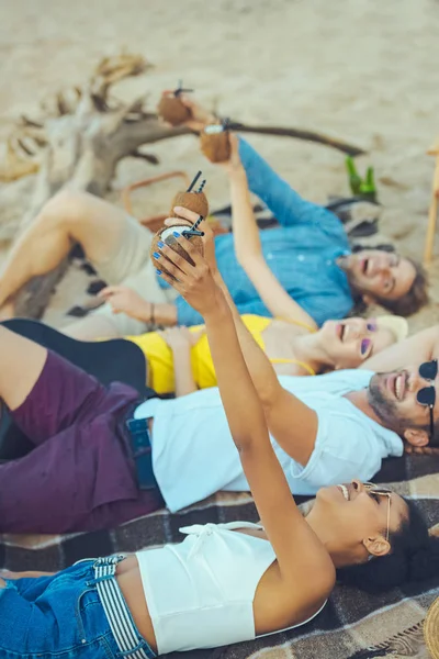 Vista lateral de amigos multiculturales con cócteles de coco descansando en la playa de arena - foto de stock