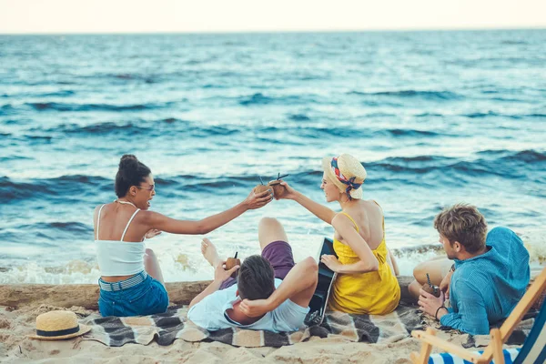 Мультикультурные друзья с кокосовыми коктейлями и акустической гитарой отдыхающие на песчаном пляже — стоковое фото