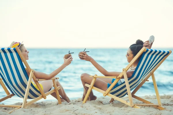 Young multiethnic women with coconut cocktails resting in beach chairs by sea — Stock Photo