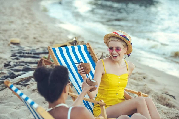 Mulheres multiétnicas jovens com coquetéis de coco descansando em cadeiras de praia por mar — Fotografia de Stock