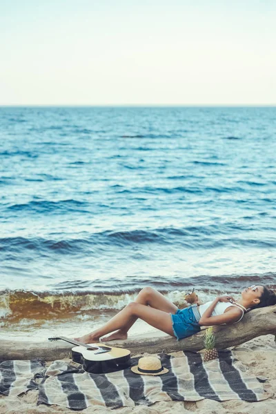 Jovem afro-americana com bebida de coquetel descansando em tronco de madeira na praia — Fotografia de Stock