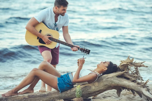 Hombre tocando la guitarra acústica para la novia afroamericana con coctel de coco en la playa de arena - foto de stock
