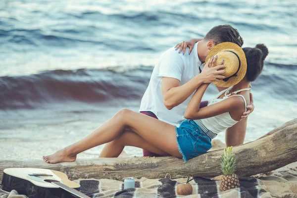 Obscured view of multiethnic couple hiding behind hat while kissing on beach — Stock Photo