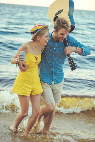 Happy couple with soda drink and acoustic guitar walking on beach — Stock Photo