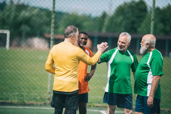 Interracial elderly football players shaking hands after match on green field — Stock Photo