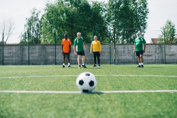 Selective focus of football ball and multicultural elderly men on green football field — Stock Photo