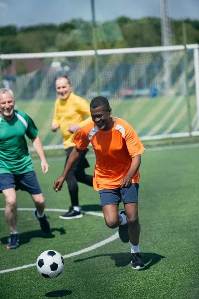 Vieil homme multiracial jouant au football ensemble sur un terrain vert — Photo de stock