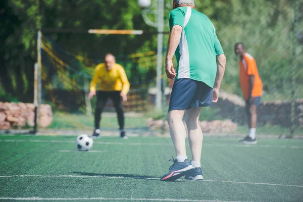 Vieil homme multiracial jouant au football ensemble sur un terrain vert — Photo de stock