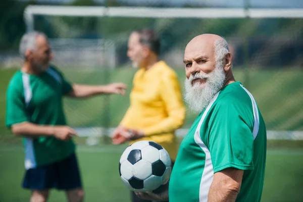 Foco seletivo do velho homem barbudo com bola de futebol e amigos atrás em campo — Fotografia de Stock