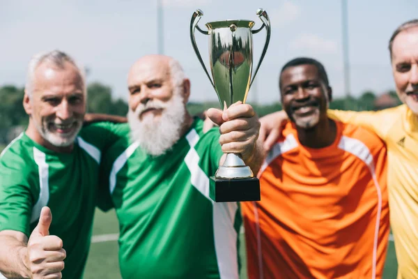 Enfoque selectivo de los viejos deportistas sonrientes multiétnicos con la copa de campeones de pie en el campo de fútbol - foto de stock