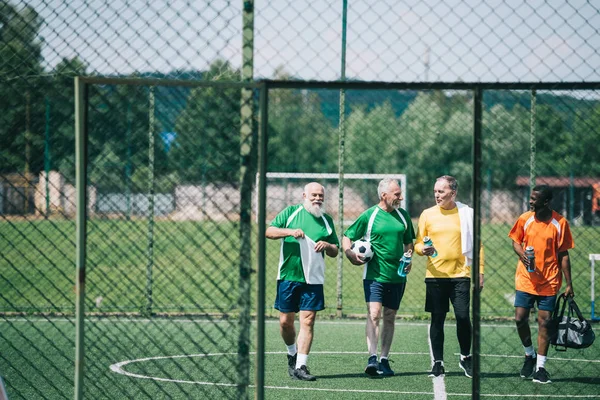 Group of interracial elderly sportsmen with sportive water bottles walking on football field — Stock Photo