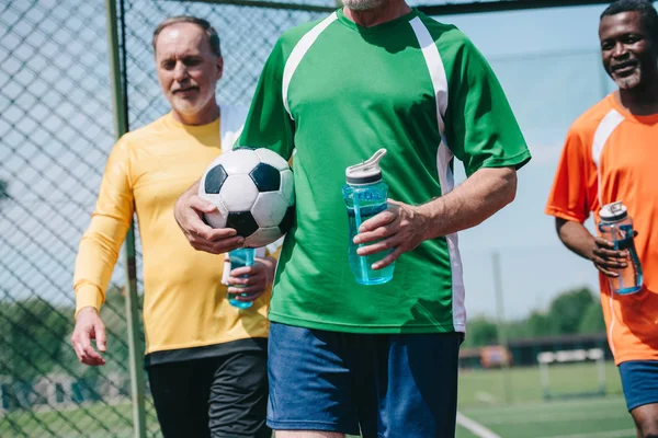 Partial view of multicultural elderly men with sportive water bottles and football ball — Stock Photo