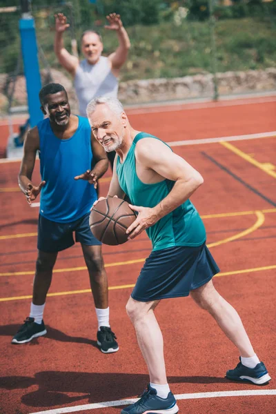 Вибірковий фокус старого з баскетбольним м'ячем в руках і багаторасовими друзями позаду на ігровому майданчику — стокове фото