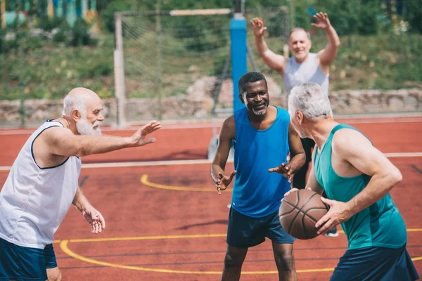 Interracial ancianos deportistas jugando baloncesto juntos en patio de recreo — Stock Photo