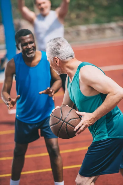 Hombres ancianos multirraciales jugando baloncesto juntos en el patio de recreo en el día de verano - foto de stock