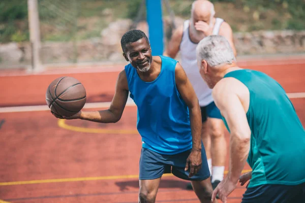 Homens idosos multirraciais jogando basquete juntos no playground no dia de verão — Fotografia de Stock