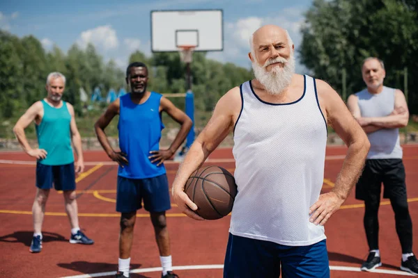 Selective focus of multiethnic elderly sportsmen with basketball ball on playground — Stock Photo