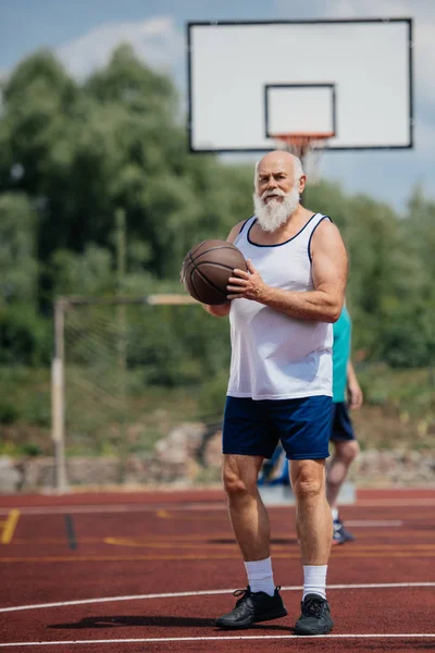 Selective focus of elderly bearded man with basketball ball in hands on playground — Stock Photo