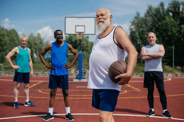 Selective focus of multiethnic elderly sportsmen with basketball ball on playground — Stock Photo