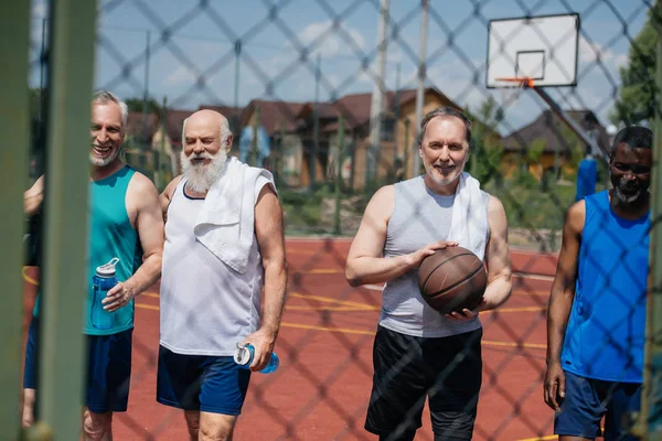 Sonrientes deportistas multiétnicos de edad avanzada con pelota de baloncesto en el patio - foto de stock