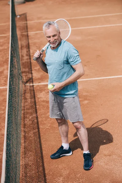Uomo anziano sorridente in abbigliamento sportivo con racchetta da tennis e palla in piedi sul campo — Foto stock