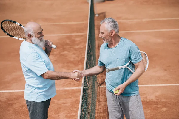 Side view of smiling elderly friends with tennis racquets shaking hands after game on court — Stock Photo