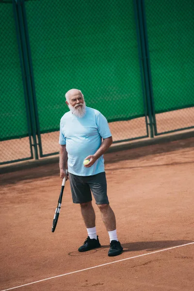 Пожилой человек в спортивной форме с теннисной ракеткой и мячом на корте — стоковое фото