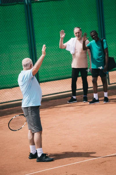 Multiethnic old men greeting friend with tennis racquet on court — Stock Photo
